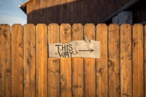 Sign on a wooden fence with an arrow pointing right and labeled ‘This Way’.